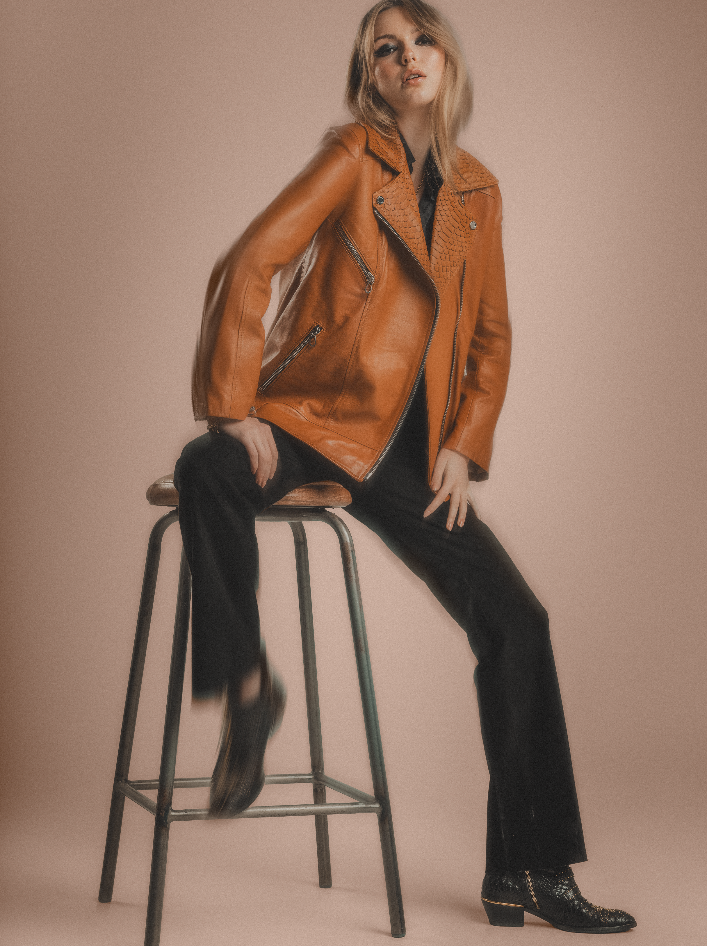 Woman wearing a brown leather jacket and black pants sitting on a stool against a beige background